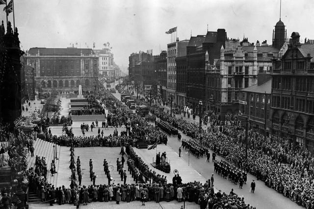 This view shows crowds on The Headrow, outside the Town Hall and on Victoria Gardens. A march past is taking place, led by the Navy, followed by military vehicles and personnel reaching down The Headrow as far as the eye can see.