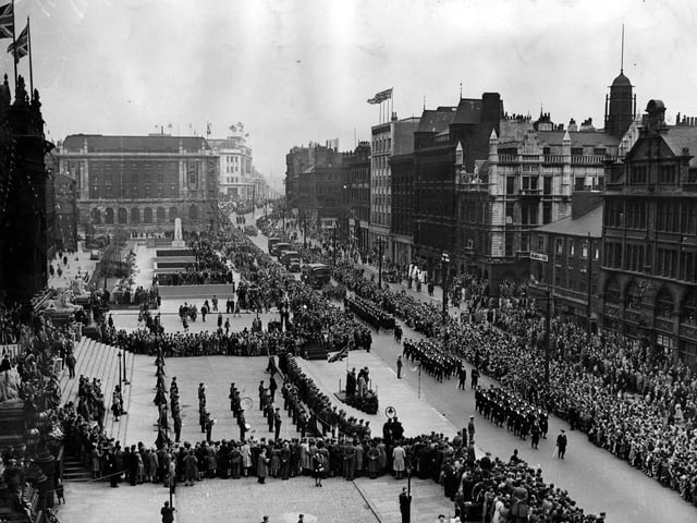 This view shows crowds on The Headrow, outside the Town Hall and on Victoria Gardens. A march past is taking place, led by the Navy, followed by military vehicles and personnel reaching down The Headrow as far as the eye can see.