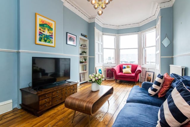 The living room has ornate cornice-work and a ceiling rose, an open shelved press, original sanded and varnished floorboards, and a deep bay window offering plenty of natural light.