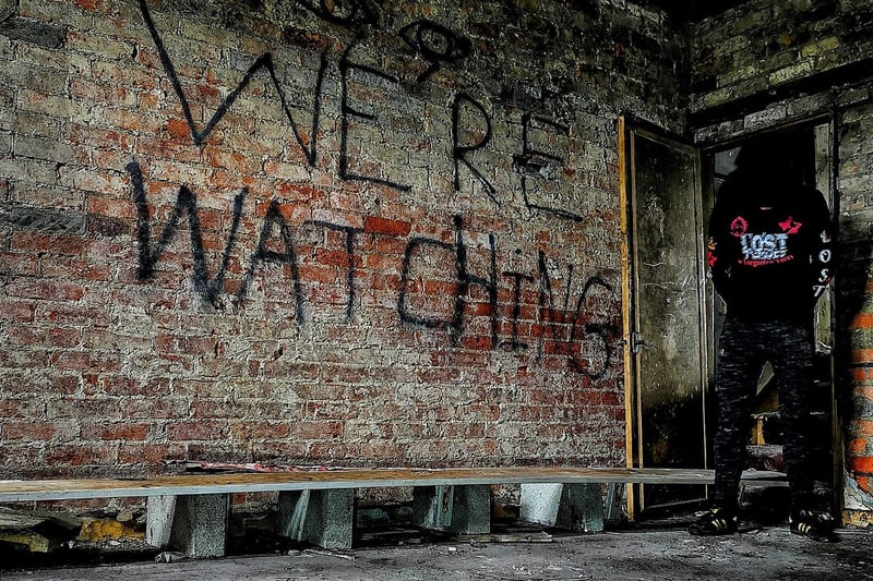 Urban explorer Lost Places & Forgotten Faces inside a room with 'We're Watching' graffitied on the wall.